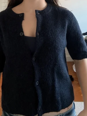 Svart kortärmad kofta från Arket - En svart kofta med korta ärmar och rund hals. Koftan är stickad i ett mjukt material och har fem knappar framtill. Perfekt att slänga över ett linne eller t-shirt. Inga defekter och använts ett fåtal gånger. 