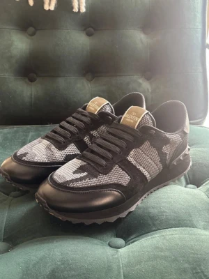 Valentino Garavani svarta camo sneakers - Snygga svarta sneakers från Valentino Garavani med coolt camo-mönster i mesh och mocka. Skorna har svarta läderdetaljer, klassiska snörningar och en robust sula med nitar på hälen. Perfekt för dig som vill ha en clean och fräsch look. Storlek 43 men passar det som har 42 också!
