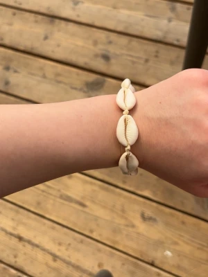 Armband med snäckor och pärlor, köpt i Spanien - Säljer ett armband med vita snäckor och små pärlor, perfekt för dig som gillar strandvibbar och naturliga material. Armbandet har en enkel och stilren design med snäckorna trädda på en beige tråd. Ingen synlig märkning.