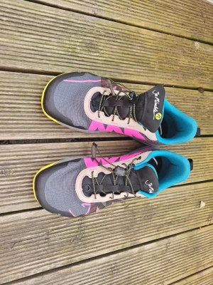 Gråa sneakers från Joe Nimble - Snygga sneakers från Joe Nimble med grå bas, rosa detaljer och beige partier. Skorna har svarta snören, turkos innersida och gula sulor med grovt mönster. Materialet är syntet och mesh, perfekt för dig som gillar färgstarka och sportiga skor.