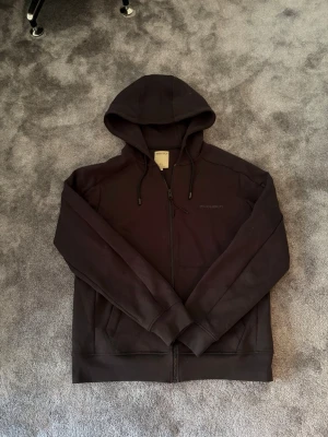 Svart hoodie med dragkedja från Woolrich - Säljer en svart hoodie från Woolrich med dragkedja framtill och två fickor. Hoodien har justerbar huva med snören och diskret logga på bröstet. Tillverkad i mjukt material.