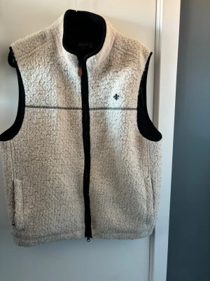 Beige fleeceväst från Morris - Säljer en beige fleeceväst från Morris med svart dragkedja och detaljer. Västen har hög krage, två fickor och en liten broderad logga på bröstet. Materialet är mjukt och fluffigt, perfekt för lager på lager och en chill look. Nypris ligger på ca 1000 kr. Hör av er vid intresse!