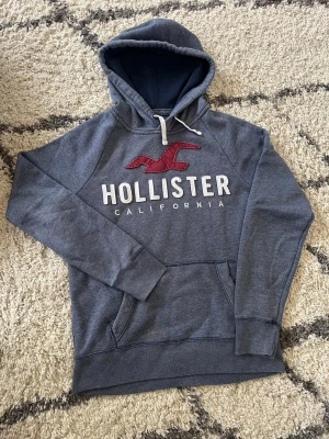 Mörk blå hoodie från Hollister - Snygg grå hoodie från Hollister med stor röd logga. Tillverkad i mjukt sweatshirtmaterial, perfekt för chill dagar, hoodien är storlek S men passar mer som en M. Skriv för mer info eller mer bilder, pris är diskuterbart!