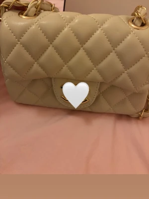 Beige quiltad handväska med kedja - Stilren beige handväska i quiltat mönster med guldfärgade detaljer och kedjerem med inslag av beige läder. Klassisk rektangulär form och vridlås framtill. Perfekt accessoar för att lyfta din outfit.