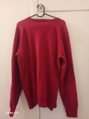 Röd stickad tröja med liten logga - Röd stickad tröja med V-ringning och lång ärm. Tröjan har en liten broderad logga på bröstet och ribbstickade muddar vid ärmslut och nederkant. Perfekt för lager-på-lager och enkel att matcha med jeans eller kjol.