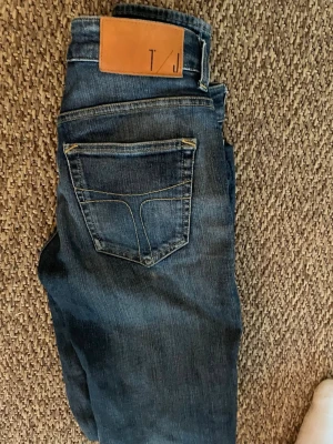 Blå jeans från T/J med läderpatch - Säljer ett par blå jeans från T/J med klassisk femficksdesign och snygg läderpatch bak. Jeansen har en slim fit och är gjorda i denim med coola slitningar och kontrastsömmar. Perfekta för dig som gillar en avslappnad och trendig look.