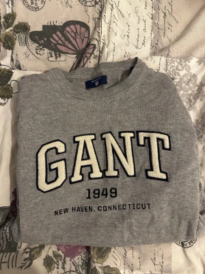 Grå sweatshirt från GANT - Klassisk grå sweatshirt från GANT med stor vit logga och texten '1949 New Haven, Connecticut' broderat på bröstet. Tröjan har rund halsringning och ribbade muddar. Perfekt för en avslappnad och stilren look.