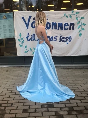 Ljusblå långklänning med öppen rygg - Säljer en elegant ljusblå långklänning i satin med smala axelband och öppen rygg med snörningsdetalj. Klänningen har en svepande kjol och en stilren, figurnära överdel. Perfekt för bal eller bröllop! Det står ingen storlek och eftersom det är snörning kan nog en xxs-s ha den :) inte använd på många år och hoppas att den ska kunna få ett nytt kärleksfullt hem 🌟🌸