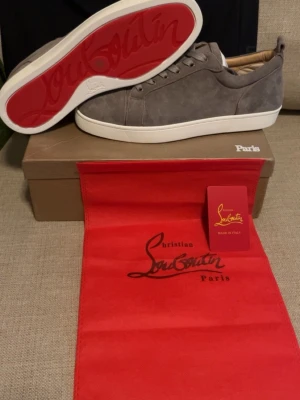 Gråa sneakers från Christian Louboutin - Säljer ett par gråa sneakers från Christian Louboutin med klassisk röd undersida och vita sulor. Skorna är låga och har en stilren design i mocka med matchande gråa skosnören. Perfekt för dig som vill ha något exklusivt och trendigt.