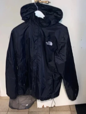 Svart vindjacka från The North Face - Svart vindjacka från The North Face med huva och dragkedja framtill. Jackan har en klassisk logga på bröstet och är tillverkad i ett lätt, vattenavvisande material. Perfekt för blåsiga dagar och enkel att packa ner. Passar S för killar och L för tjejer. Snöret där nere har åkt ut men går att sätta in igen