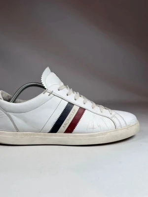 Moncler sneakers  - Snygga vita sneakers från Moncler med klassiska röda och marinblå ränder på sidan. Skorna har lågt skaft, vita snören och Moncler-logga på plösen. Tillverkade i läder för en clean och stilren look.
