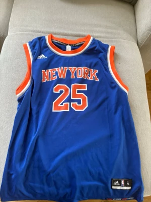 New York Knicks basketlinne #25 Rose 2017 Jersey  - Blått basketlinne från Adidas med New York Knicks tryck och nummer 25 på fram- och baksidan. Namnet Rose står på ryggen. Linnet har orange och vita detaljer runt ärmhål och hals. Officiell NBA-produkt i lätt och ventilerande material.