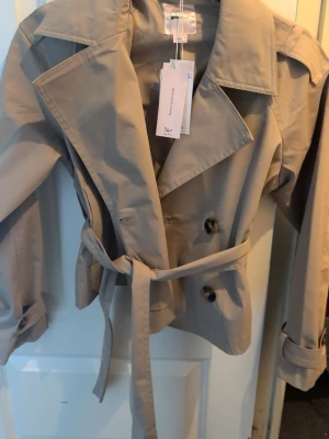 Kort beige trenchcoat med bälte - Stilren kort trenchcoat i beige från Gina tricot. 