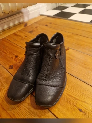 Svarta stövletter från Rieker(37) - Säljer ett par eleganta svarta ankelboots från Rieker i storlek 37. Skorna är endast använda en enda gång.  Varför köpa dessa?  Nyskick: Inget slitage på sulan eller skinnet – de ser ut som de precis kommit ur kartongen.  Rieker Antistress: Otroligt lätta och bekväma. Perfekta om du går eller står mycket under dagen.  Detaljer: Fin dekorativ blomma och smidig dragkedja på insidan.  Kvalitet: Rejäl sko som andas och håller fötterna glada.   