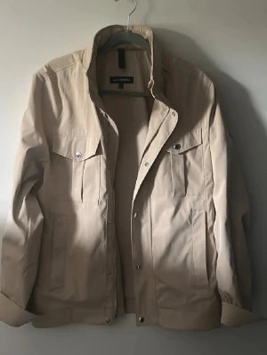 J.Lindeberg vit fieldjacka - Stilren beige overshirt från J.Lindeberg med klassisk krage, tryckknappar och dragkedja. Två bröstfickor med lock och två sidofickor. Perfekt lager för en clean och modern look. Materialet är slätt och lätt, passar till många outfits.