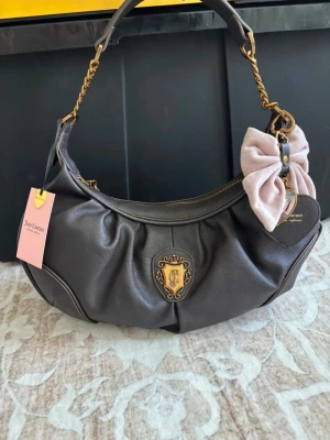 Juicy Couture brun axelväska med rosett - Mörkbrun axelväska från Juicy Couture i mjukt konstläder med guldfärgade detaljer och kedja. Väskan har en stor ljusrosa sammetsrosett och dekorativt JC-emblem framtill. Perfekt för dig som gillar en söt och trendig look.