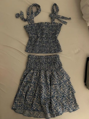 Blommigt blått set med volangkjol - Säljer ett superfint tvådelat set med smockad topp och volangkjol.Setet är köp på en marknad för 300kr. Setet är blått med små vita och gula blommor, toppen har knytbara axelband och kjolen har flera volanger. Perfekt för sommaren och riktigt söt stil!