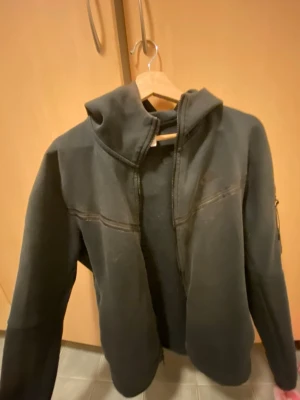 Svart hoodie från Nike med dragkedja - Svart hoodie från Nike med dragkedja framtill och huva. Tröjan har diskret Nike-logga på bröstet och en ficka på ärmen. Perfekt för en sportig och avslappnad stil. Nike tech/ lite solskadad!
