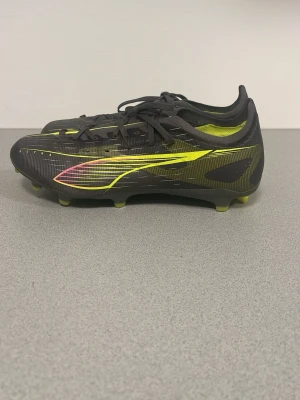 Puma svarta och gula fotbollsskor - Säljer ett par snygga fotbollsskor från Puma i svart med gula och neonrosa detaljer. Skorna har låg profil, snörning och mönstrad ovandel i syntetmaterial för bra grepp på planen. Perfekta för dig som vill sticka ut på matchen!
