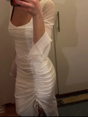 Vit studentklänning  - Säljer en vit studentklänning med transparenta trekvartsärmar och snygg snörning framtill. Klänningen har en tight passform och rynkade detaljer längs sidorna för en extra stilren look. Perfekt för dig som gillar trendiga och feminina plagg.