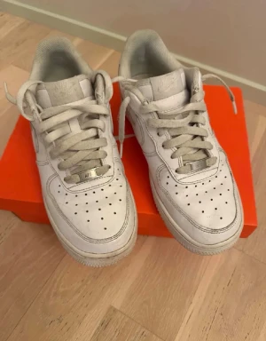 Nike Air Force 1 vita sneakers - Klassiska Nike Air Force 1 sneakers i helvitt skinn med snörning och perforerade detaljer på tån. Ikonisk design med Nike-logga på sidan och stabil sula. Perfekta för en stilren och sportig look.