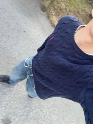 Mörkblå kabelstickad tröja Polo Ralph Lauren - Mörkblå kabelstickad tröja från Polo Ralph Lauren med rund halsringning och röd broderad logotyp på bröstet. Tröjan har ribbstickade muddar vid ärmslut och nederkant. Perfekt för en stilren och klassisk look.