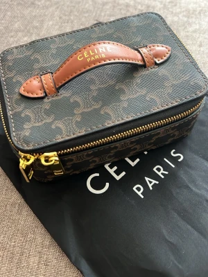  C Paris brun monogram handväska - Snygg liten handväska från C Paris i brunt monogrammönstrat canvasmaterial med detaljer i ljusbrunt eco skinn. Väskan har guldfärgad dragkedja och ett stilrent handtag upptill. Perfekt för dig som gillar exklusiva accessoarer med klassisk design. Storlek S