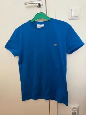 Blå t-shirt från Lacoste - Snygg blå t-shirt från Lacoste med klassisk rund hals och det ikoniska krokodilmärket broderat på bröstet. T-shirten är kortärmad och tillverkad i mjuk bomull, perfekt för en stilren och avslappnad look. Pris kan diskuteras 
