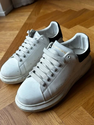 Alexander McQueen vita sneakers - Säljer ett par vita sneakers från Alexander McQueen med svart detalj på hälen och guldfärgad logga. Skorna har tjock sula, klassisk låg modell och är tillverkade i läder. Perfekt för dig som vill ha en stilren och trendig look. Skorna är lite smutsiga men är fortfarande i bra skick och därför väljer jag att sänka priset, går att tvätta.