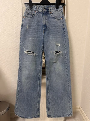Ljusa raka jeans med slitningar - Säljer ett par ljusblå jeans med raka ben och trendiga slitningar på båda benen. Jeansen har hög midja, klassiska fem fickor och stängs med knapp och dragkedja. Perfekta för en avslappnad och cool stil.