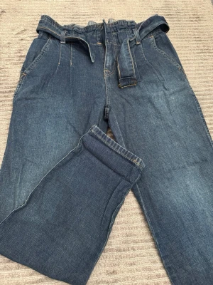 Mörkblå paperbag jeans med skärp - Säljer ett par mörkblå paperbag jeans med hög midja och tillhörande denimskärp. Jeansen har raka ben, två framfickor och två stora bakfickor. Materialet är klassisk denim och passformen är avslappnad och trendig.