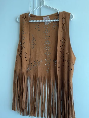 Brun mockaimitationsväst med fransar - Cool brun väst i mockaimitation med laserskurna mönster och långa fransar nedtill. Perfekt för en boho-inspirerad look. Ärmlös och öppen framtill, ger en avslappnad och trendig stil.
