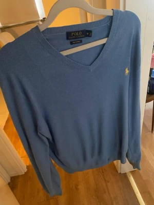 Blå stickad tröja från Polo Ralph Lauren - Snygg blå stickad tröja från Polo Ralph Lauren med V-ringning och klassisk gul broderad logga på bröstet. Tröjan har långa ärmar och ribbade muddar vid ärmslut och nederkant. Perfekt för lager-på-lager och chill stil.