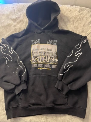 Svart hoodie The Couture Club - Säljer en svart hoodie från The Couture Club med stor huva och tryck framtill och baktill. Framsidan har texten 'FEAR LESS LOVE MORE' och 'COUTURE', medan baksidan har tour-datum och 'WORLDWIDE TOUR'. Ärmarna har vita flames-prints för extra streetstyle.