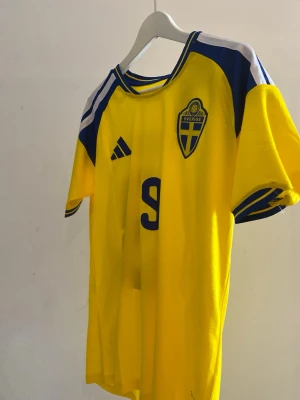 Sverige fotbollströja Isak #9 Adidas - Gul och blå officiell Sverige fotbollströja från Adidas med nummer 9 och namnet Isak på ryggen. Tröjan har korta ärmar, rund hals och Sveriges landslagsemblem på bröstet. Perfekt för dig som vill stötta landslaget på match och speciellt nu under en stundande vm💛💙