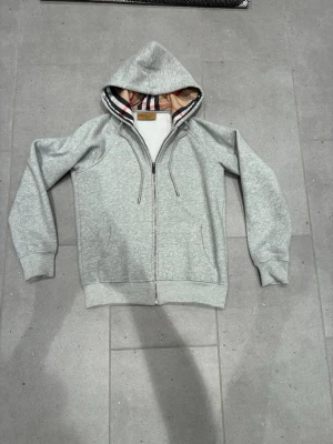 Grå hoodie från Burberry - Säljer en grå hoodie från Burberry med dragkedja och klassiskt rutigt foder i huvan. Hoodien har broderad logga på bröstet och ribbade muddar. Perfekt för dig som vill ha en stilren och exklusiv look. Materialet är mjukt och bekvämt.