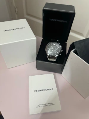 Emporio Armani silverfärgad klocka svart  - Stilren klocka från Emporio Armani med silverfärgat armband i metall och svart urtavla. Klockan har kronograf-funktion, tydliga index och logga upptill. Levereras i originalask med tillhörande manual och garanti.