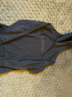 Mörkblå hoodie från Calvin Klein - Snygg mörkblå hoodie från Calvin Klein med klassisk logga på bröstet. Hoodien har huva med dragsko och en stor magficka framtill. Perfekt för en chill och stilren look. Tillverkad i mjukt material som känns skönt mot huden.