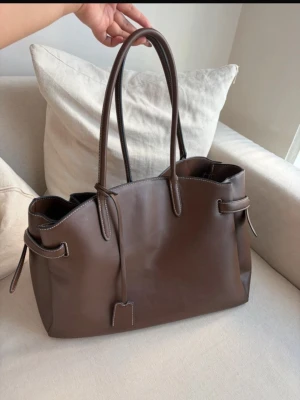 Brun tote handväska i läderlook - Stilren brun handväska i slät läderimitation med långa handtag och dekorativa knytband på sidorna. Väskan har en rymlig design och kontrastsömmar som ger en snygg detalj. Perfekt för dig som vill ha en klassisk och trendig accessoar till din outfit.