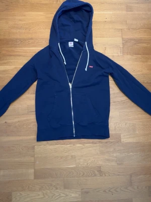 Marinblå zip hoodie från Levi's - Säljer en marinblå hoodie från Levi's med dragkedja framtill och vita snören i huvan. Klassisk passform med två fickor fram och liten röd Levi's-logga på bröstet. Perfekt för en avslappnad och stilren look. Köpt i butik för 800, men har tyvärr inte kvar kvitto.