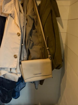 Beige axelväska från Calvin Klein - Snygg beige axelväska från Calvin Klein med stilren design och justerbar rem. Väskan har ett lock med magnetstängning och flera fack inuti för enkel organisering. Tillverkad i slätt konstläder med diskret logga framtill.
