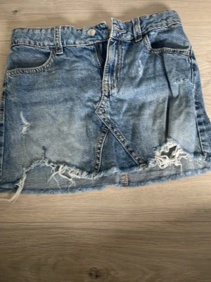 Blå jeanskjol med fransig kant - Snygg blå jeanskjol med fransig och sliten nederkant. Klassisk femficksmodell med knapp och dragkedja framtill. Perfekt för en avslappnad och trendig look.