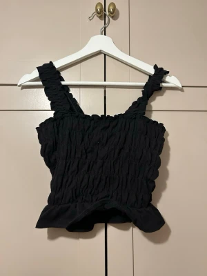 svart smockad linnetopp - Trendig svart linnetopp med smockad struktur och breda axelband. Toppen har en lätt volang nertill. aldrig använd 