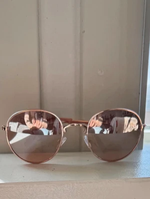 Runda rosa solglasögon i metall - Snygga solglasögon med rund form och tunna bågar i roséguld. Glasen är lätt tonade i rosa och bågarna har en stilren metallfinish med rosa detaljer på skalmarna. Perfekta för att lyfta din look med en trendig touch.