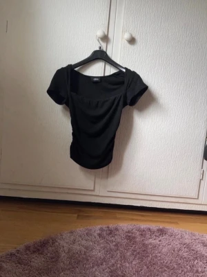 Svart topp med fyrkantig ringning - Snygg svart topp från Cider med korta ärmar och trendig fyrkantig ringning. Toppens passform är figurnära och materialet känns mjukt och stretchigt, perfekt till jeans eller kjol.