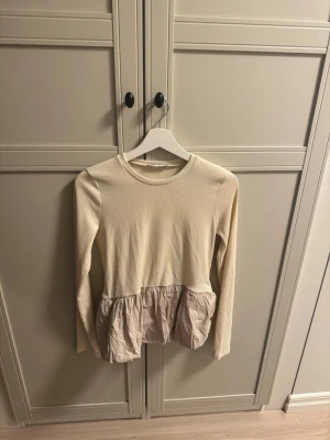 Beige långärmad topp med volang - Söt beige långärmad topp med rund halsringning och volangdetalj i ett glansigt material nedtill. Överdelen är ribbad och har en feminin, lös passform. Perfekt för dig som gillar en romantisk och trendig stil.