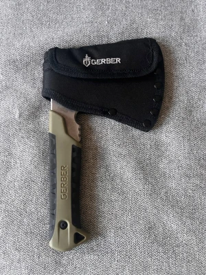 Gerber yxa med svart fodral - Kompakt äkta yxa från Gerber med svart och olivgrönt handtag i plast och gummi för bra grepp. Yxan har ett svart skyddsfodral i tyg med Gerber-logga och bältesfäste på baksidan. Perfekt för friluftsliv och camping.