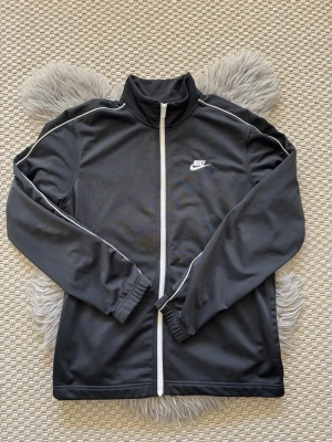Svart träningsjacka från Nike - Svart träningsjacka från Nike med vit dragkedja och vita detaljer längs ärmarna. Jackan har hög krage, elastiska muddar och Nike-logga på bröstet. Perfekt för träning eller sportiga outfits.