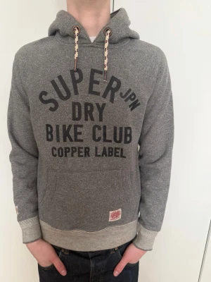 Grå Superdry hoodie med tryck - Snygg grå hoodie från Superdry med svart texttryck på bröstet och detaljerade snören i huvan. Mjuk borstad insida, känguruficka framtill och ribbade muddar vid ärmar och midja. Perfekt för en avslappnad streetwear-look.
