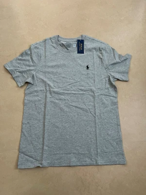 Ralph lauren T-shirt grå storlek medium  - Ny Ralph Lauren t-shirt med klassisk passform. Perfekt basplagg till sommaren.  Oanvänd Väldigt populär modell Enkel att matcha  Skickas snabbt 📦 Rabatt vid köp av flera.